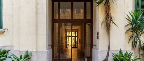 Entrada de un edificio con puerta de vidrio, plantas en macetas y un camino empedrado que conduce al interior.
