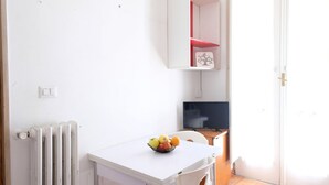 Een kleine kamer met een witte tafel, een fruitschaal, een wit kastje, een televisie en een radiator.