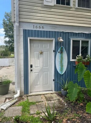 Residential building with a distinctive exterior, featuring a white door and a surfboard on the wall, surrounded by a garden and potted plants.