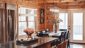 A wooden cabin interior with a kitchen island, dining table, chairs, a refrigerator, and a hanging light fixture. - The Burrow Lodge! Pet Friendly! Hot Tub! Sleeps 16 (Logan)