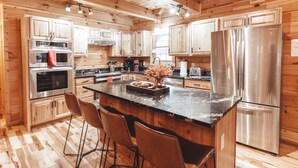 A modern kitchen with wooden cabinets, stainless steel appliances, a marble countertop, and bar stools. - The Burrow Lodge! Pet Friendly! Hot Tub! Sleeps 16 (Logan)
