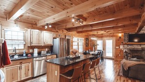 A spacious kitchen with wooden beams, a central island with bar stools, a dining area, and a stone fireplace. - The Burrow Lodge! Pet Friendly! Hot Tub! Sleeps 16 (Logan)