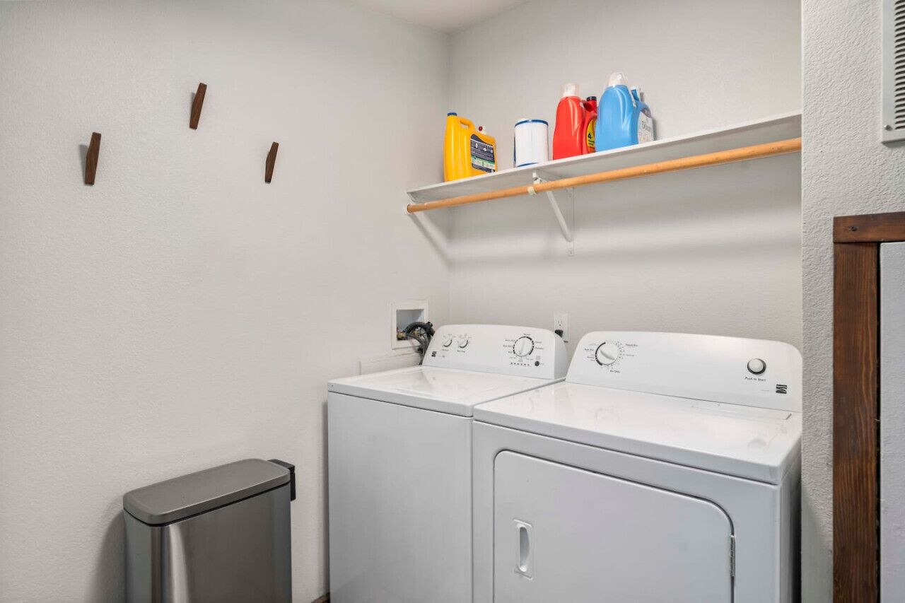 Un cuarto de lavado con dos lavadoras y secadoras blancas, un estante con productos de limpieza y un bote de basura.