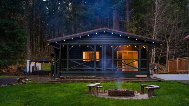 Une cabane dotée d’un porche couvert, d’un foyer et d’un petit abri de jardin, située dans une zone boisée.