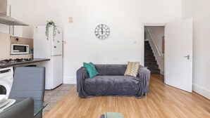 Un espace de vie compact comprenant un coin cuisine, un canapé gris, une horloge murale et un escalier menant à un autre niveau.