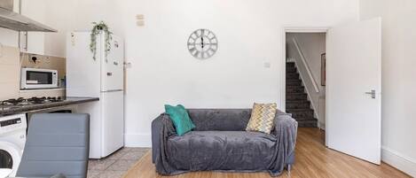 Un espacio habitable pequeño con kitchenette, un sofá gris, un reloj en la pared y una escalera que lleva a otro nivel.
