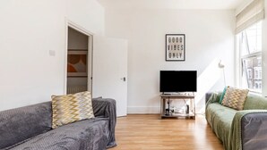 Un salon moderne avec un canapé gris, un canapé vert, un téléviseur à écran plat posé sur un meuble en bois, et un panneau mural portant l'inscription « SEULEMENT LES BONNES VIBES ».