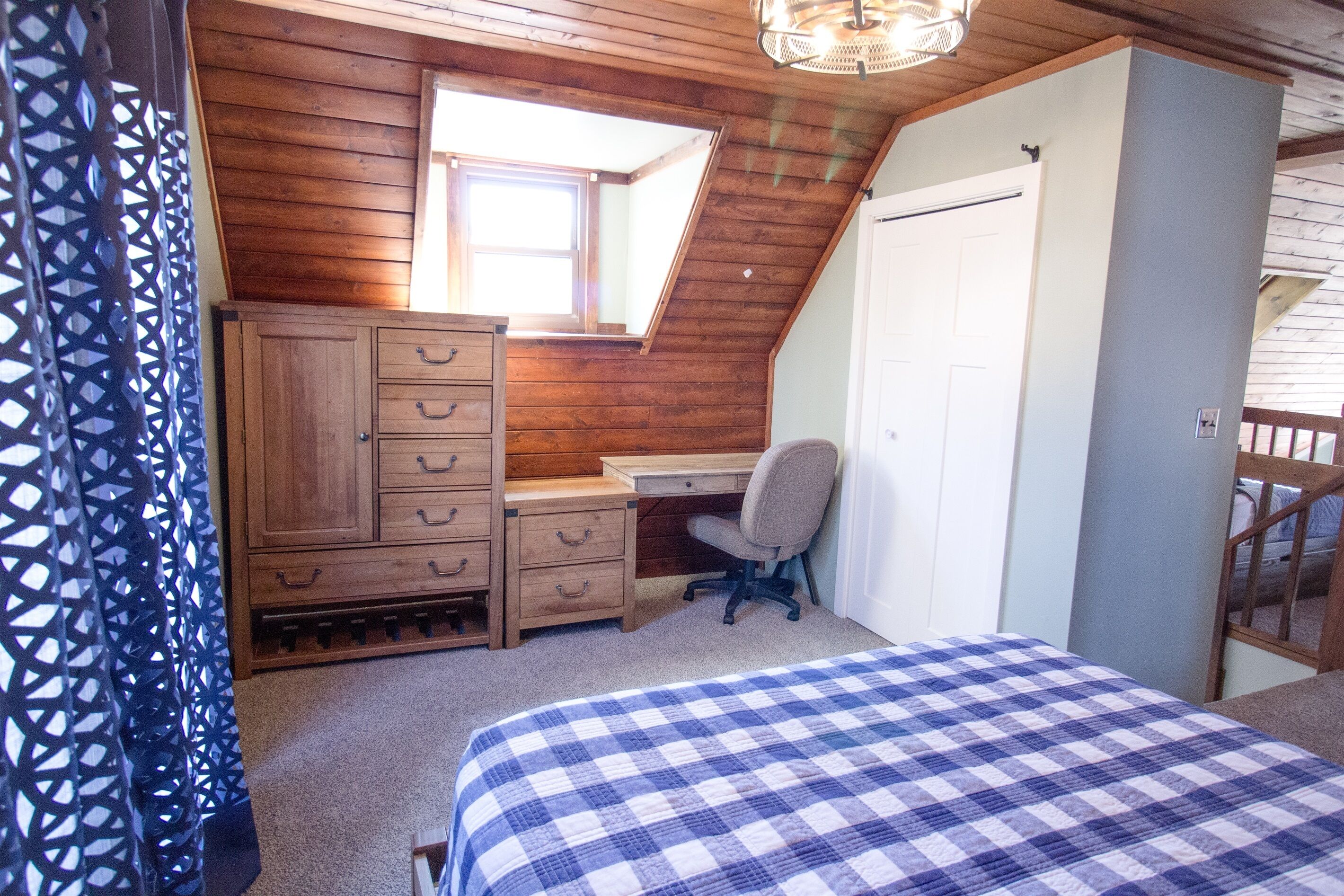 Une chambre avec un plafond en bois, un bureau avec une chaise, une armoire et un lit recouvert d’une couverture à carreaux.