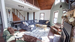 Ein geräumiges Wohnzimmer mit einer großen Eckcouch, einem Couchtisch und einem Kamin. Eine Treppe führt auf eine obere Ebene.