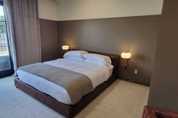 A neatly made bed with white linens and a brown throw blanket, two wall-mounted reading lamps, and a window with curtains.