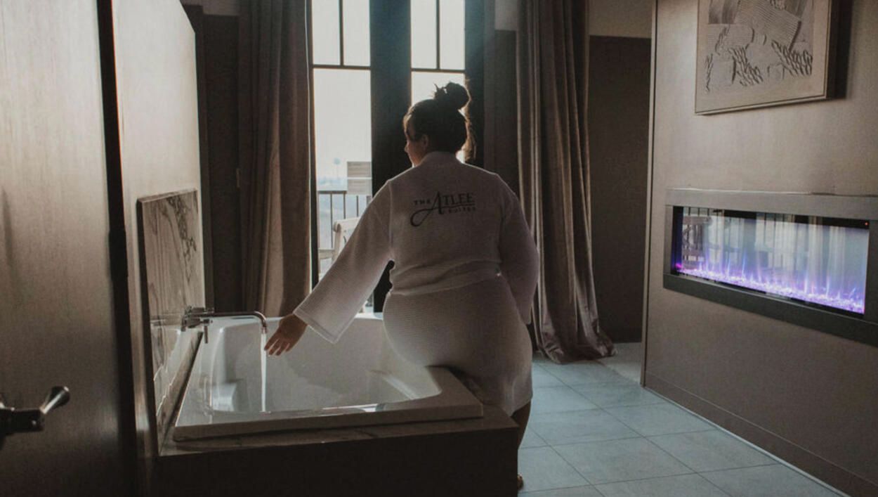 Eine Person in einem weißen Bademantel steht in einem Raum mit einer Badewanne, einem fensterbelaubten Fenster und einem an der Wand befestigten Aquarium.