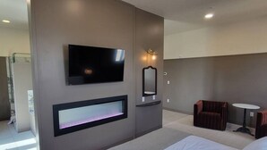 Ein modernes Hotelzimmer mit Bett, Flachbildfernseher an der Wand, integriertem Kamin, kleinem Tisch und einer Sitzecke mit Sofa und Sessel.