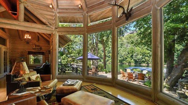 A log cabin interior with a large window overlooking a lakeside patio.