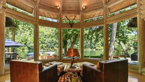 A wooden gazebo with a central table and seating area, surrounded by large windows offering a view of a lake and trees.
