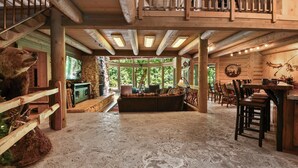 A rustic cabin interior with a stone fireplace, wooden furniture, and a large window overlooking greenery.