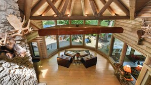 A log cabin interior with a high ceiling, wooden beams, a ceiling fan, and a large window offering a view of the outdoors.