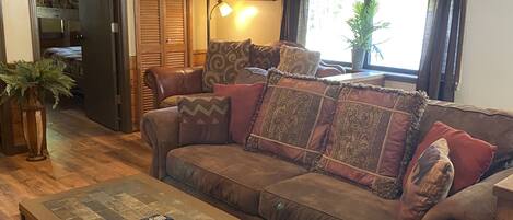 A living room with a brown sofa, patterned cushions, a glass coffee table, a lamp, and a window with a view of greenery.