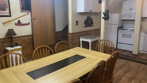 Een eetgedeelte met een houten tafel en stoelen, een kleine witte tafel met een lamp, en een keuken met witte kasten en apparatuur.