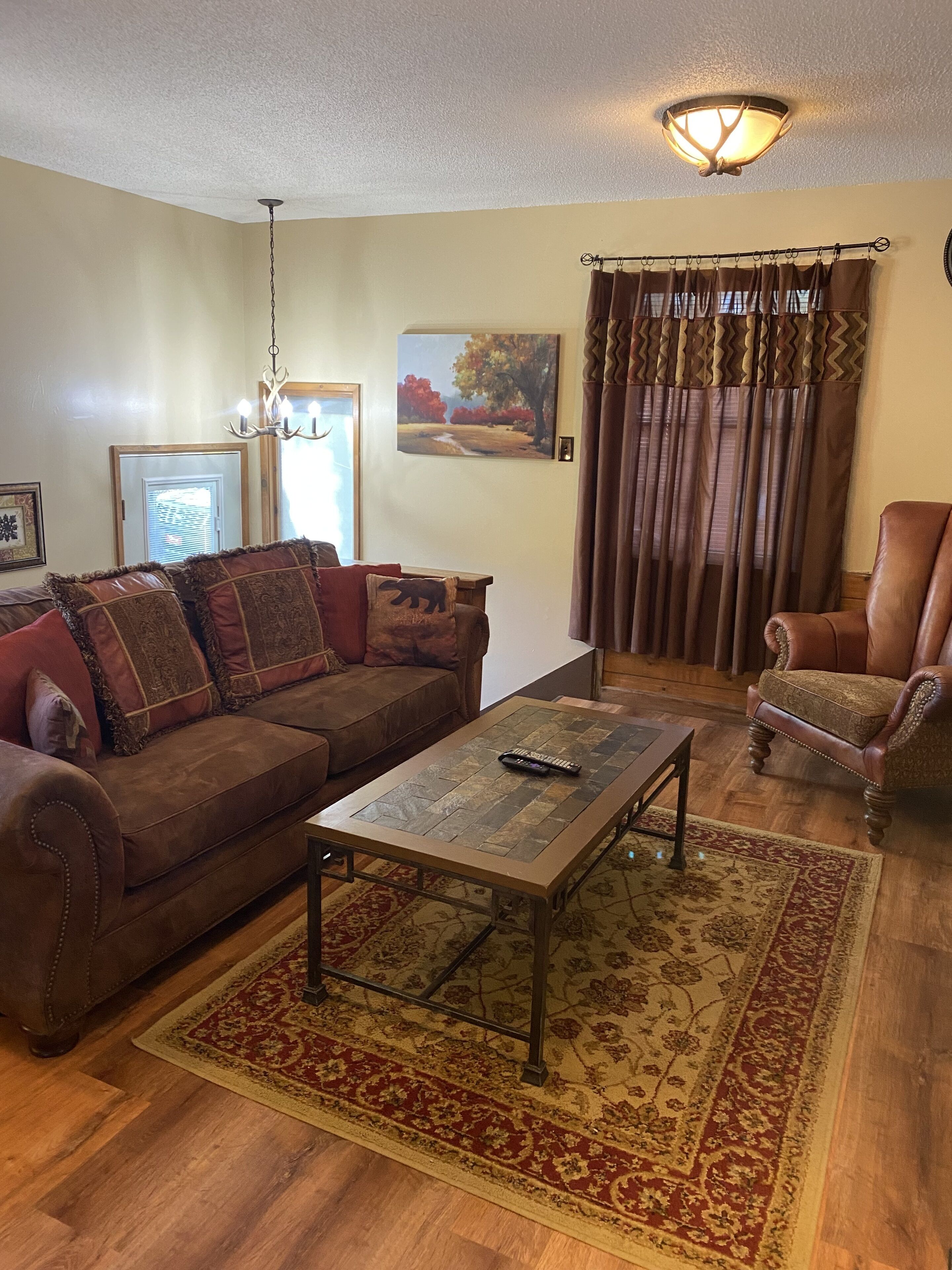 A living room with a brown leather sofa, a coffee table, a patterned rug, a wooden floor, a painting, and a window with curtains.