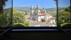 A view through a window of a building with a large, ornate church with two towers and a bell tower, surrounded by greenery and hills. - Casa das Acácias (Cabeceiras de Basto)