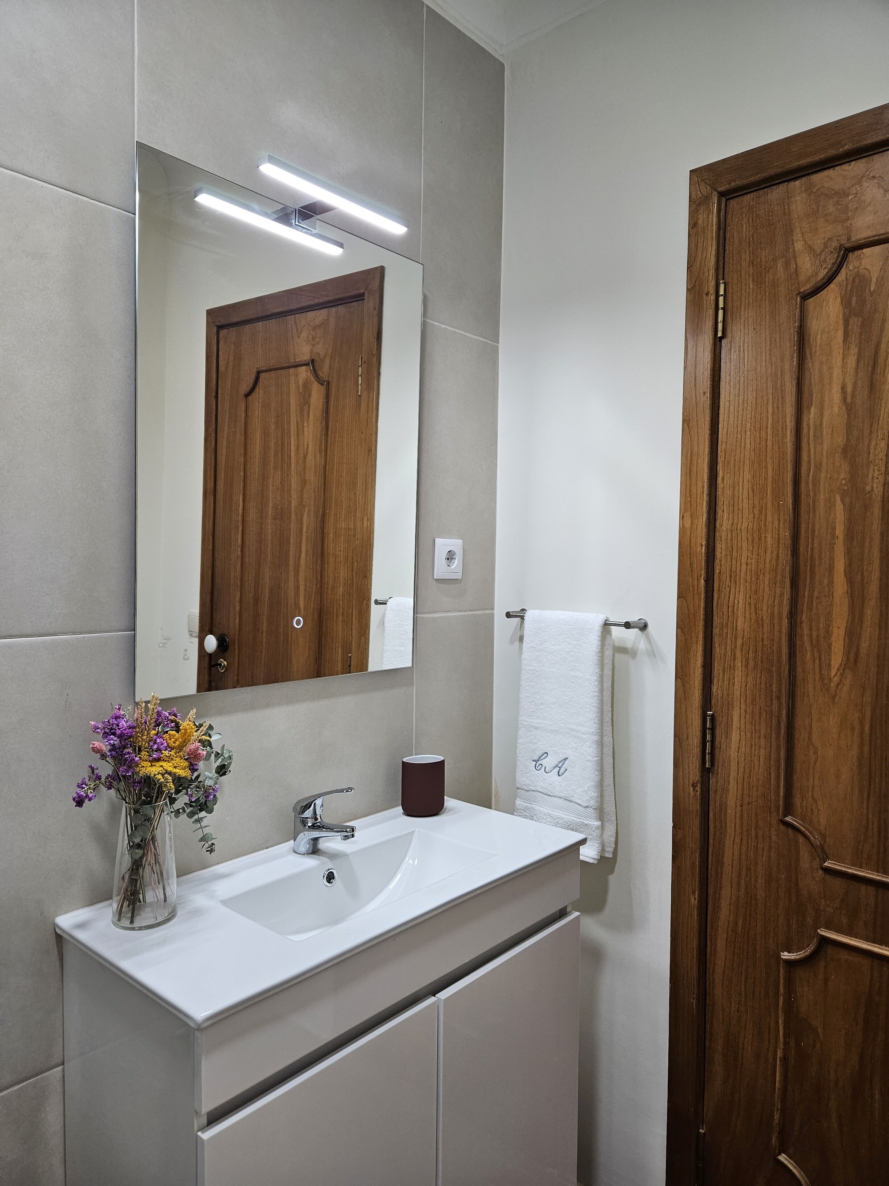 A bathroom with a wooden door, a white sink, a mirror, and a vase with flowers.