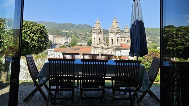 A terrace with a table and chairs, a blue umbrella, and a view of a historic building with two towers. - Casa das Acácias (Cabeceiras de Basto)