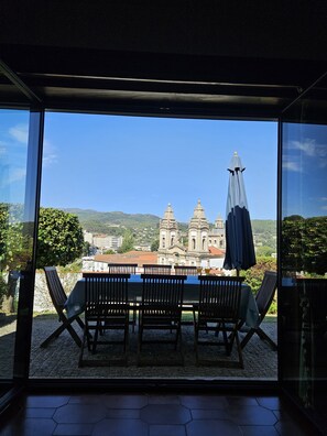 A terrace with a table and chairs, a blue umbrella, and a view of a historic building with two towers. - Casa das Acácias (Cabeceiras de Basto)