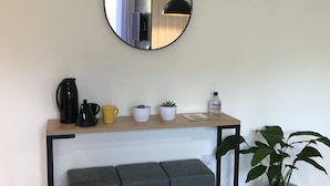 Moderni pieni keittiö, jossa on seinälle ripustettu peili, kahvinkeitin, vedenkeitin, keltainen muki ja ruukkukasvi.