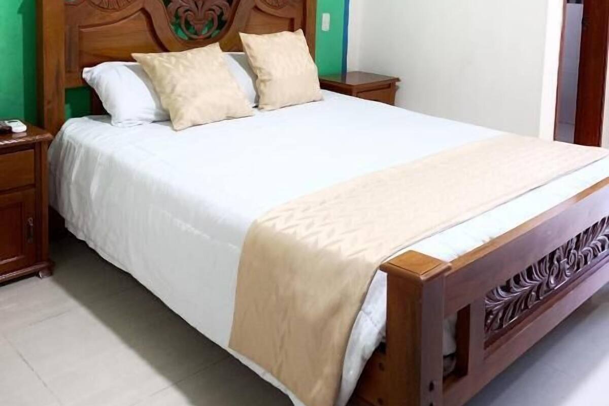 Ein Einzelbett mit Holz-Kopf- und Fußteil, einer beigen Bettdecke und zwei Kissen. Ein Nachttisch mit Lampe steht daneben. Die Wände sind grün und die Decke weiß.