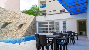 Un espace aménagé au bord de la piscine, avec un mur en pierre, une terrasse couverte et des sièges d’extérieur.