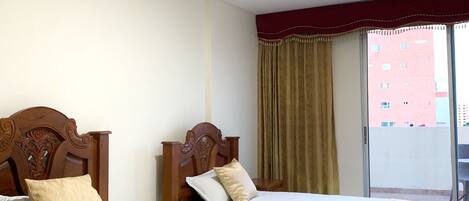 A hotel room with two beds, a wooden headboard, a window with curtains, and a view of a building outside.