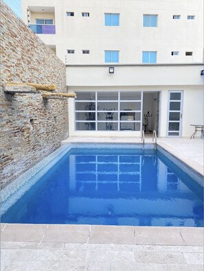 A swimming pool with a stone wall on one side and a building with multiple windows and balconies in the background. - Hotel Continental Plaza (Barranquilla)