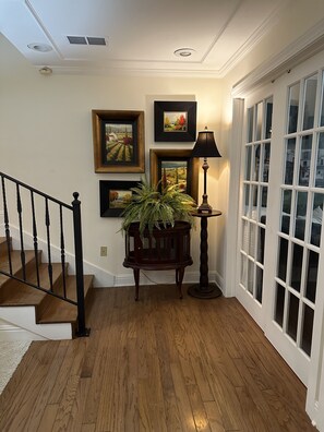 A wooden staircase with a black railing leading to a landing. There are three framed paintings on the wall above the landing. A potted fern is placed on a wooden table next to a floor lamp.