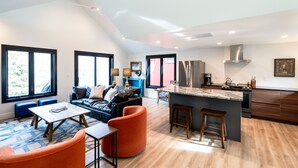 Ein modernes Wohnzimmer mit einer Couch, Hocker und einem Couchtisch. Es gibt eine Küchenzeile mit Herd, Kühlschrank und Barhocker.
