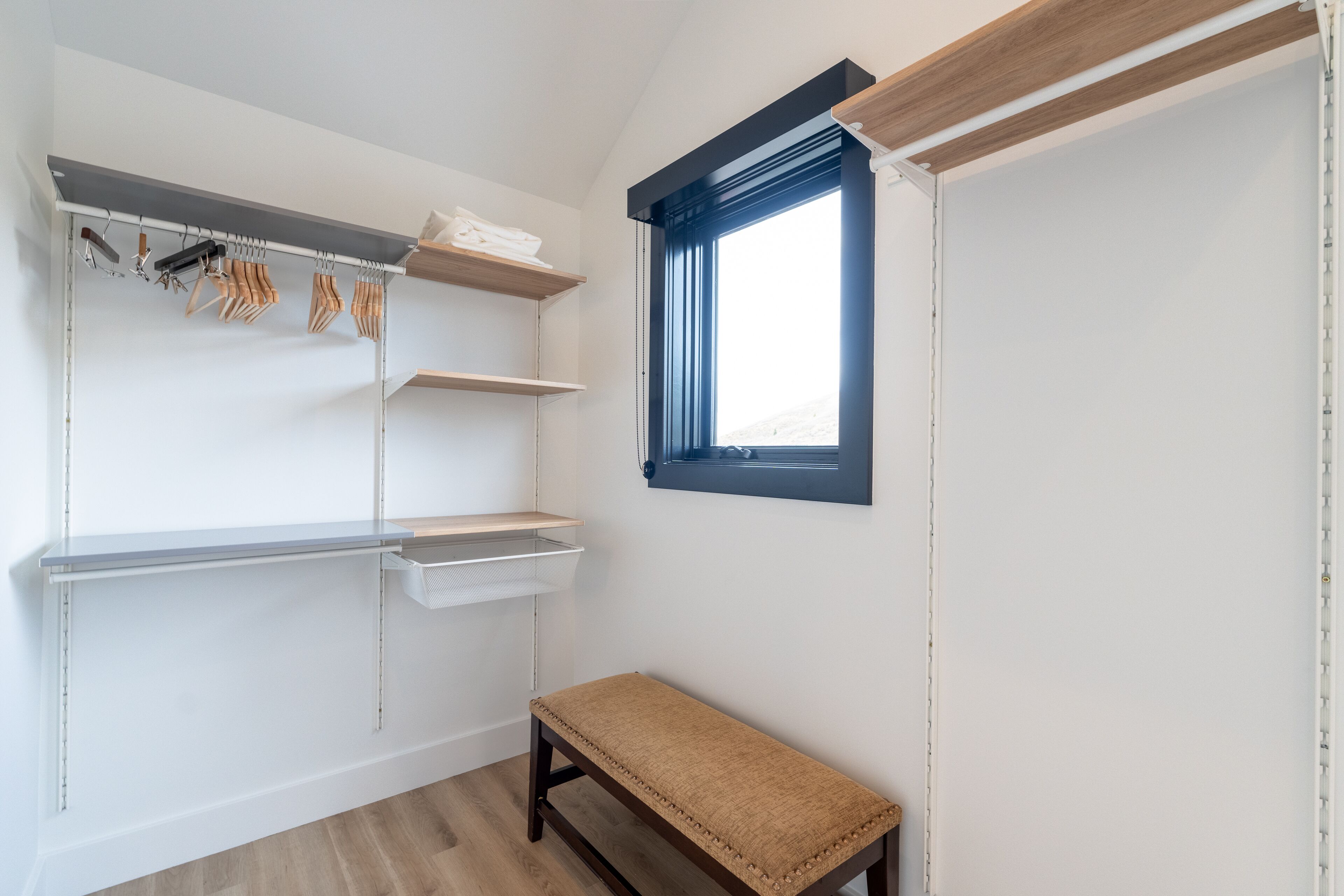 Une pièce dotée d’une banquette intégrée, d’une fenêtre avec stores et d’une étagère murale où sont rangés des vêtements et des paniers de rangement.