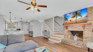 Una sala de estar moderna con chimenea, una escalera y una gran pantalla de televisión mostrando una escena de playa.