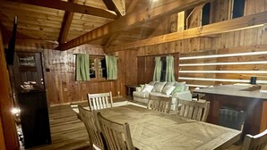 Interior de una cabaña de madera con una mesa de comedor, sillas, un sofá y una pequeña cocina.