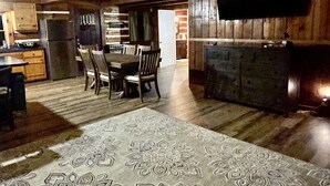 Un interior rústico con un gran tapete estampado, una mesa de comedor de madera, sillas, una zona de cocina y un televisor de pantalla plana montado en la pared.