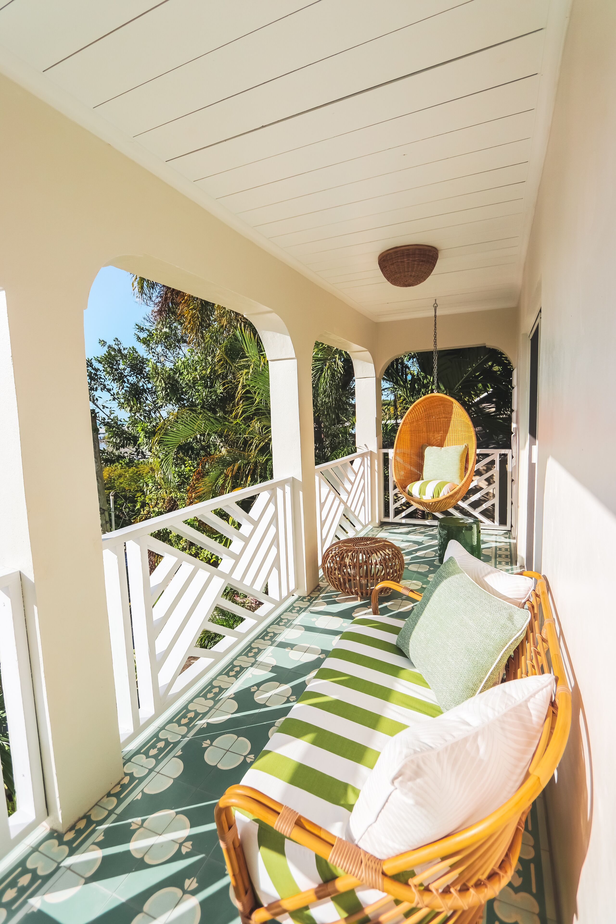 A covered porch with a wicker chair and a cushioned bench, surrounded by white railings and a patterned floor.