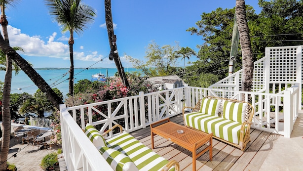 A deck with a wooden table and two striped sofas, overlooking a beach with palm trees and a white fence. - The Rock House Hotel (Dunmore Town)