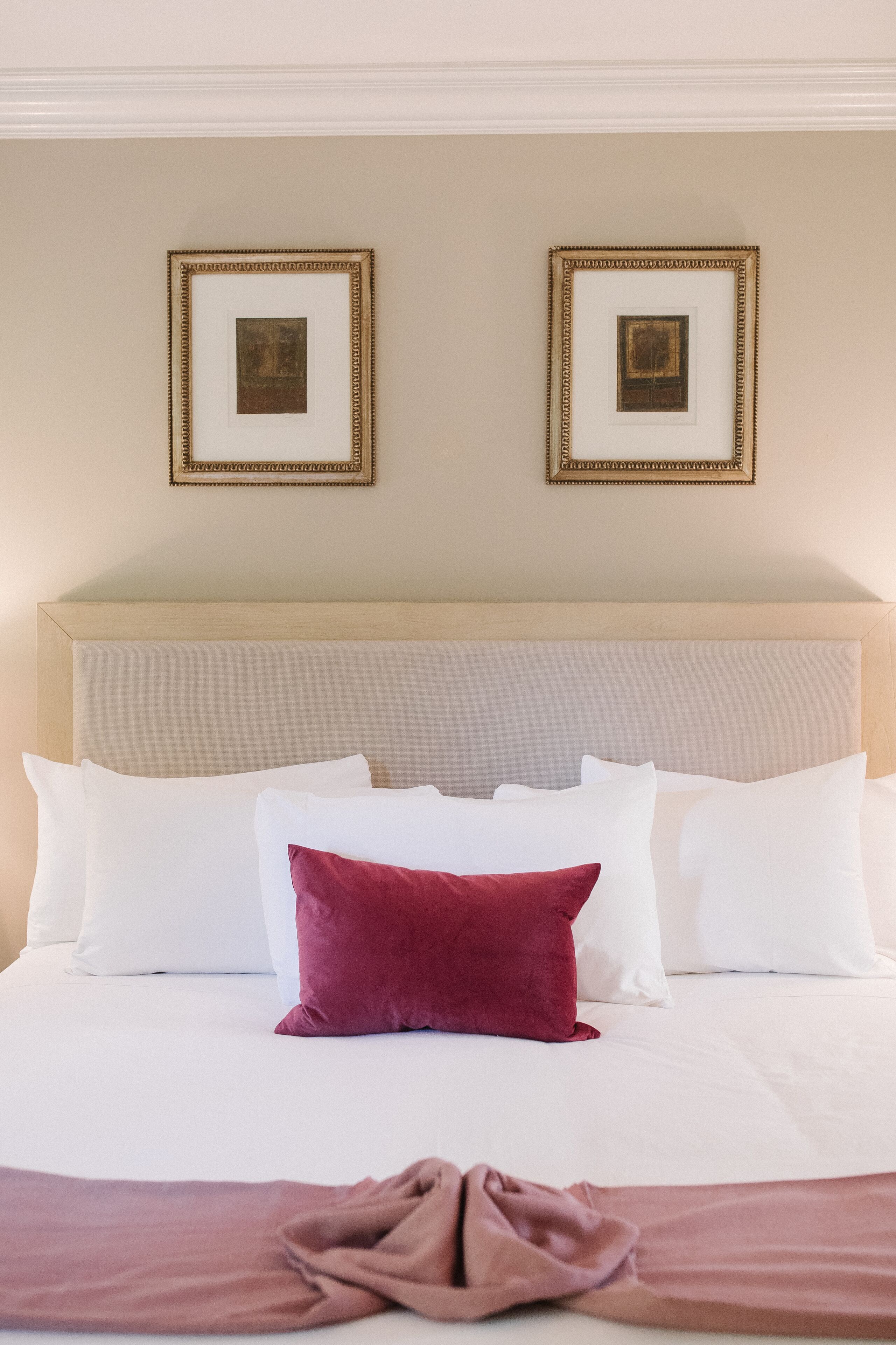 Un letto rifatto con cuscini bianchi e color bordeaux, una testiera beige e due quadri appesi al muro.
