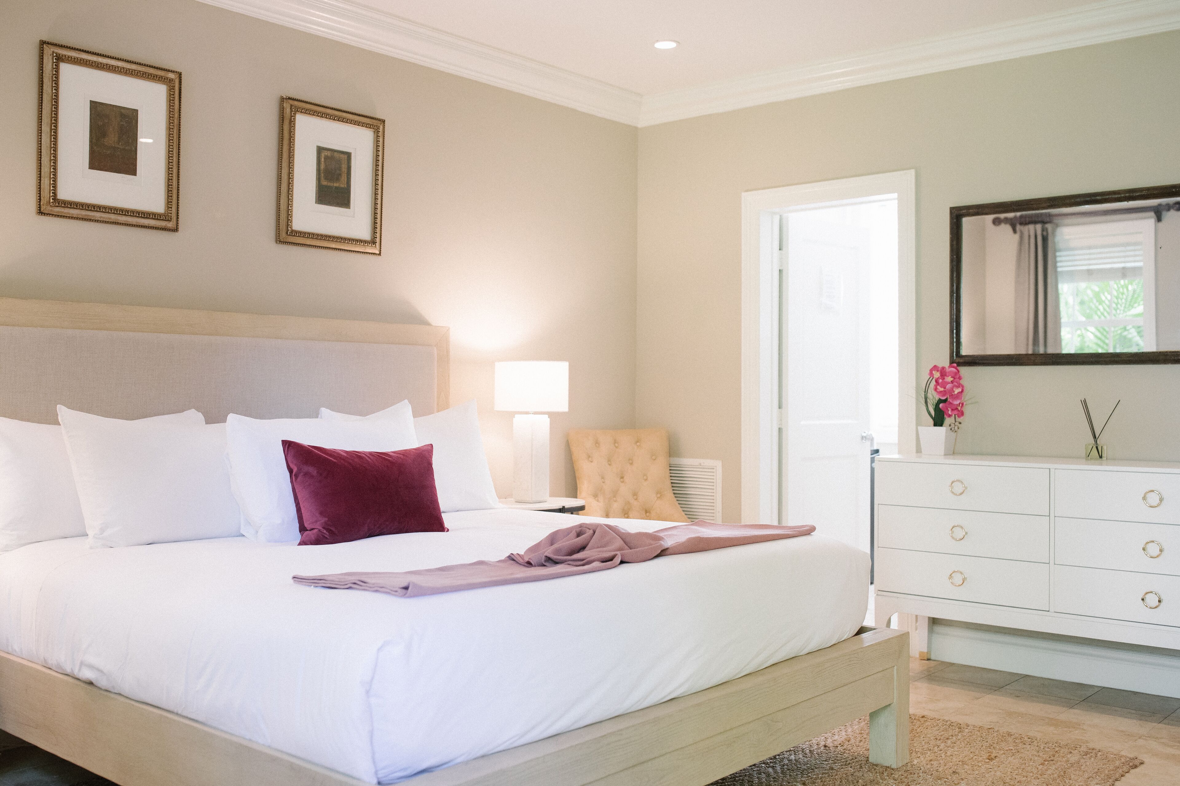 Un lit bien fait, avec des oreillers blancs et bordeaux, une tête de lit beige et une table de chevet avec une lampe. On y trouve également une commode blanche avec des tiroirs et un miroir au-dessus.