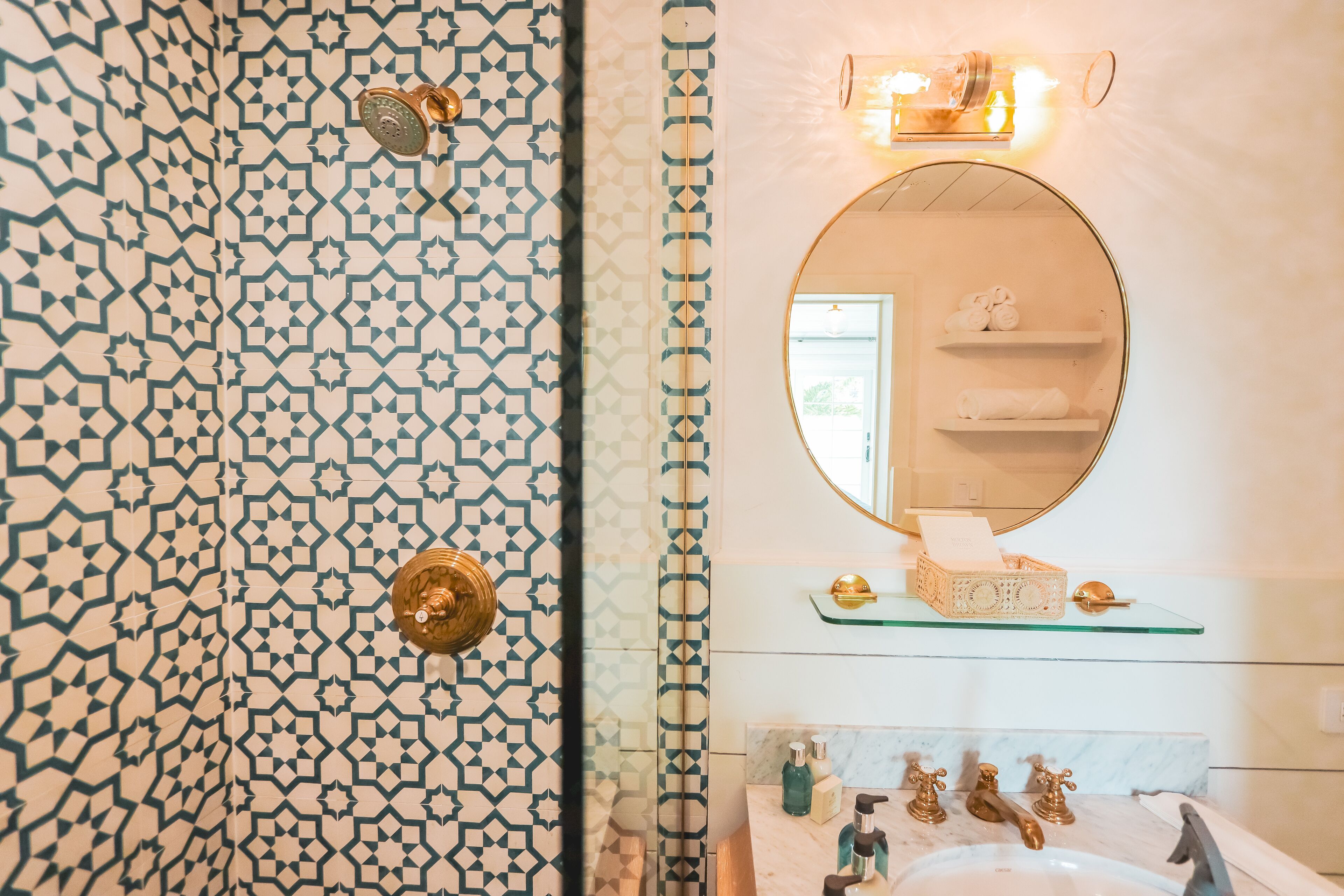 Un baño con pared de azulejos estampados, un espejo redondo, repisas de vidrio con artículos de aseo y un lavamanos de mármol.