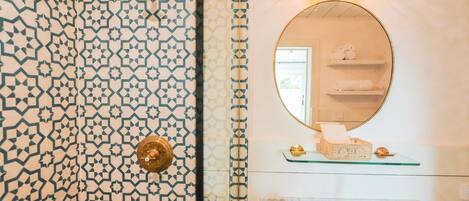 Un baño con pared de azulejos estampados, un espejo redondo, repisas de vidrio con artículos de aseo y un lavamanos de mármol.