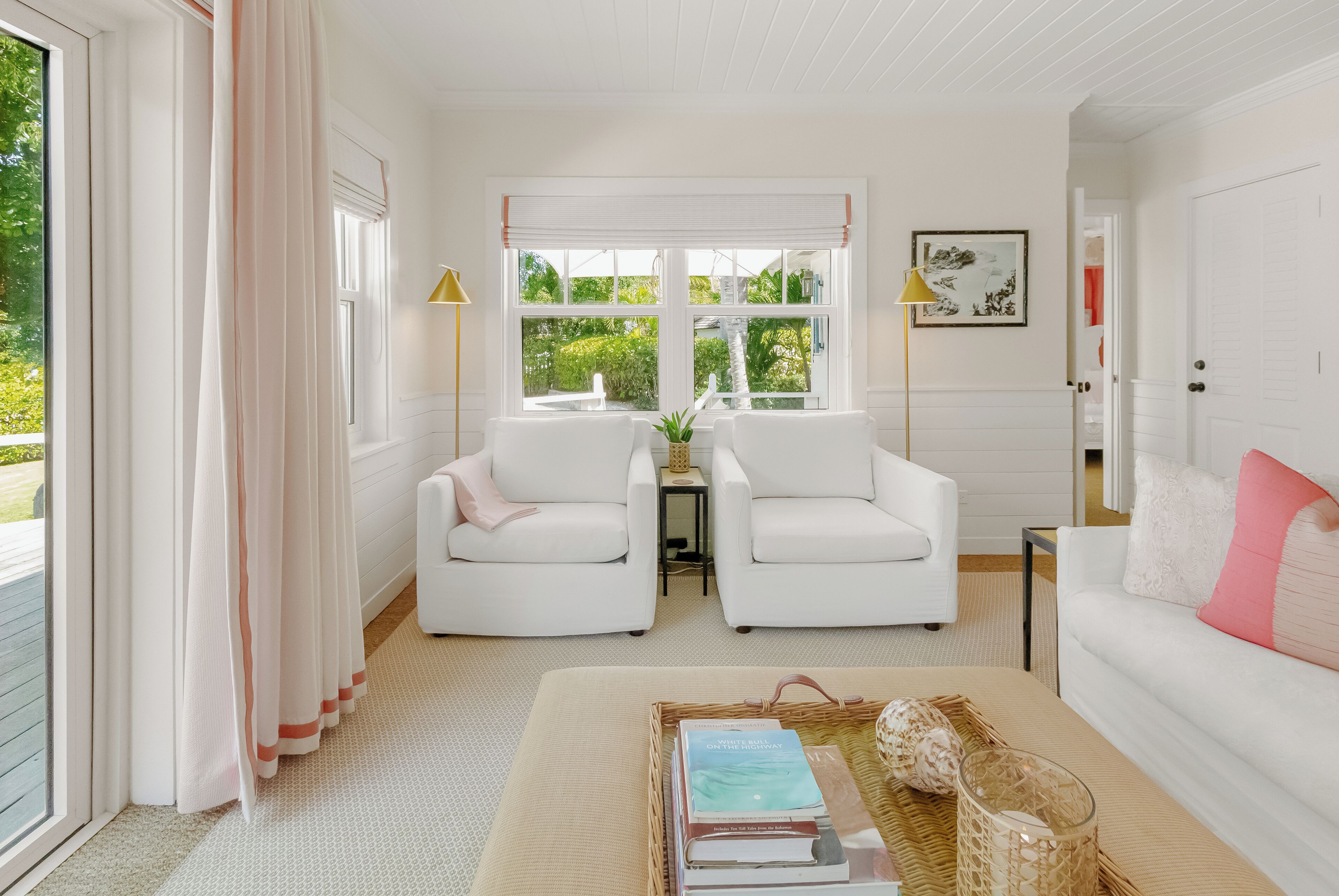A bright, white room with two white armchairs, a small table with a vase, and a large window with a view of greenery.
