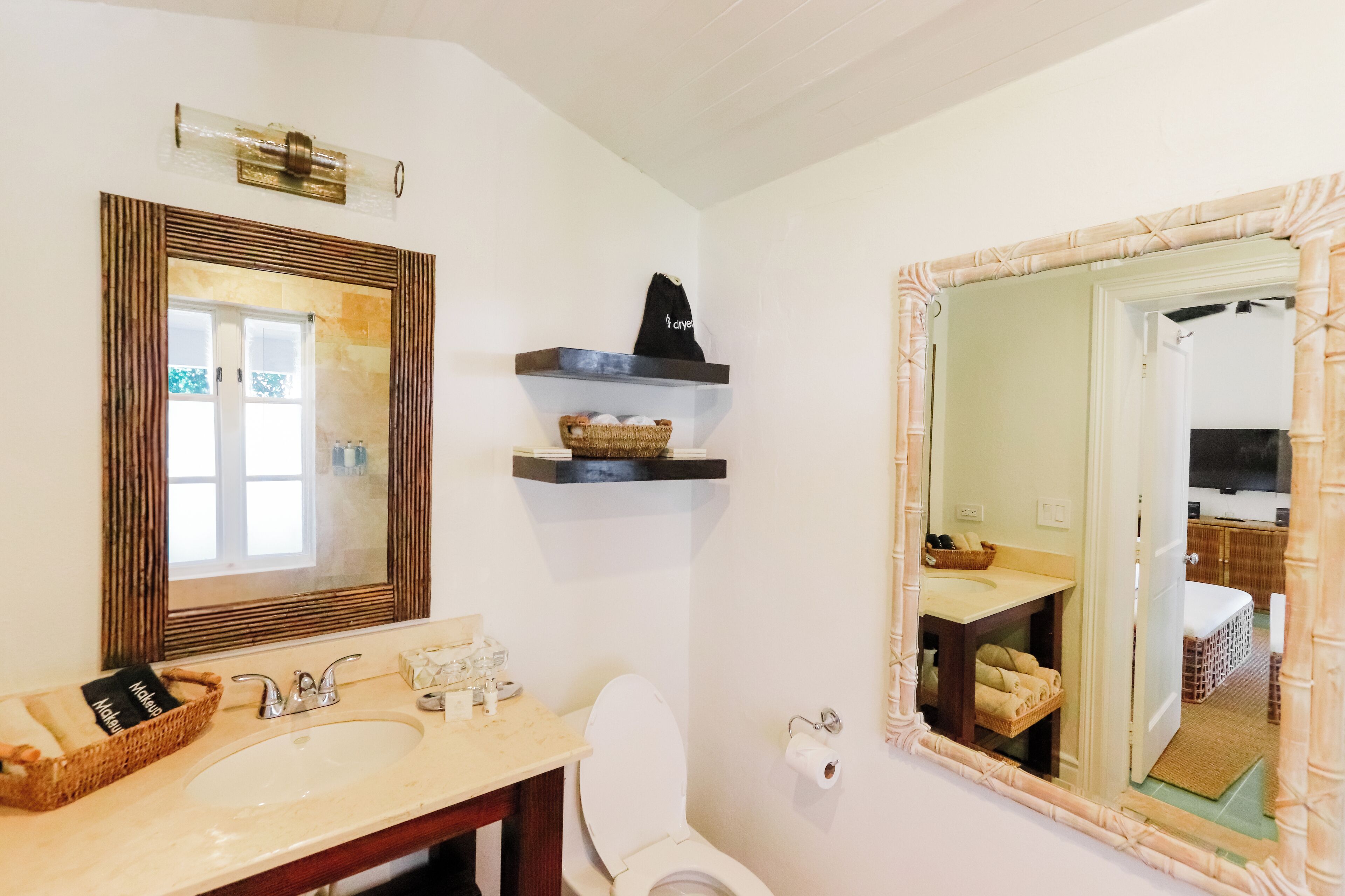 A bathroom with two sinks, a toilet, and a large mirror. There are shelves above the sinks with baskets and a towel.