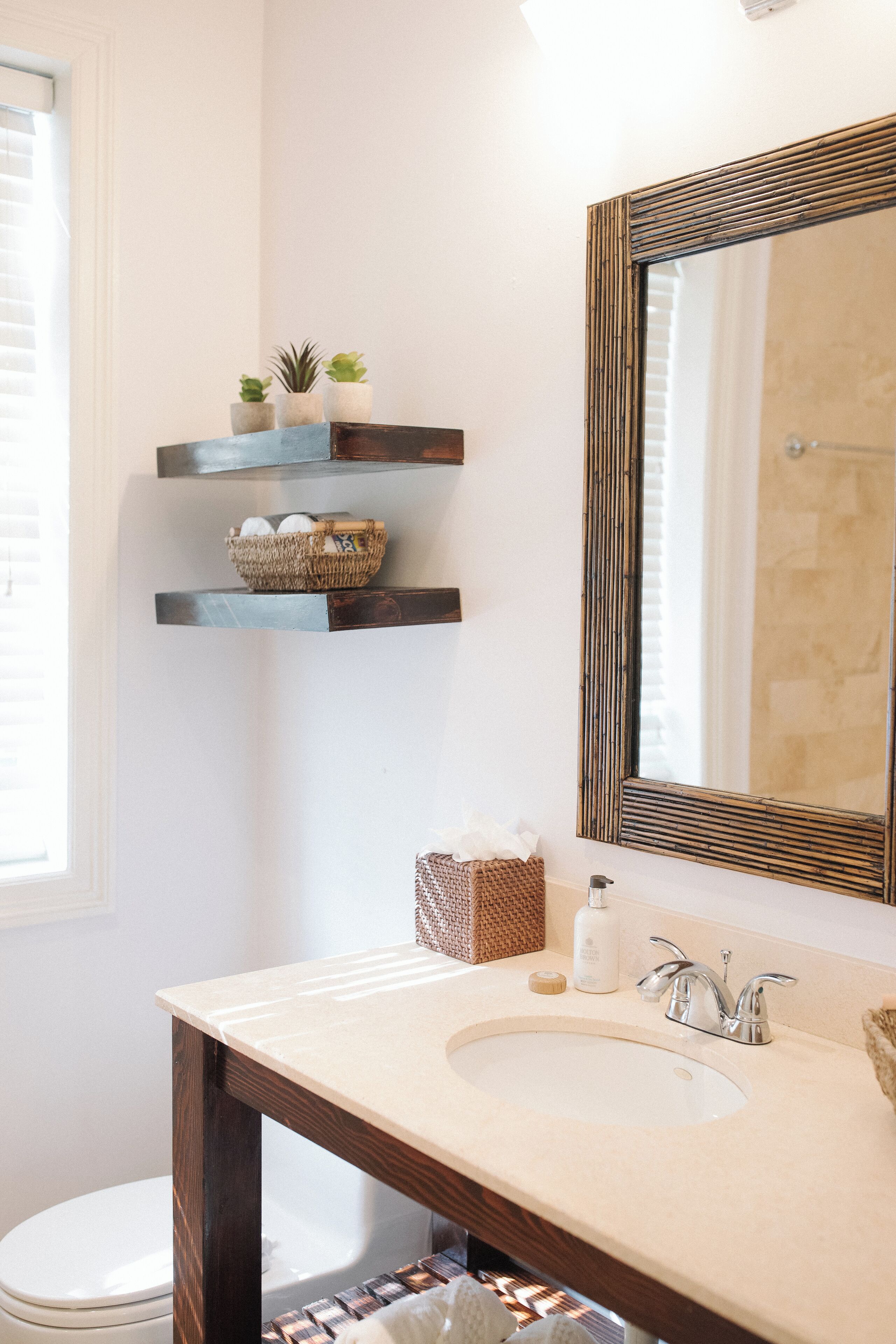 Un bagno con un lavandino bianco, un mobile contenitore in legno e un ampio specchio. Si vedono delle mensole con piante e un cesto.