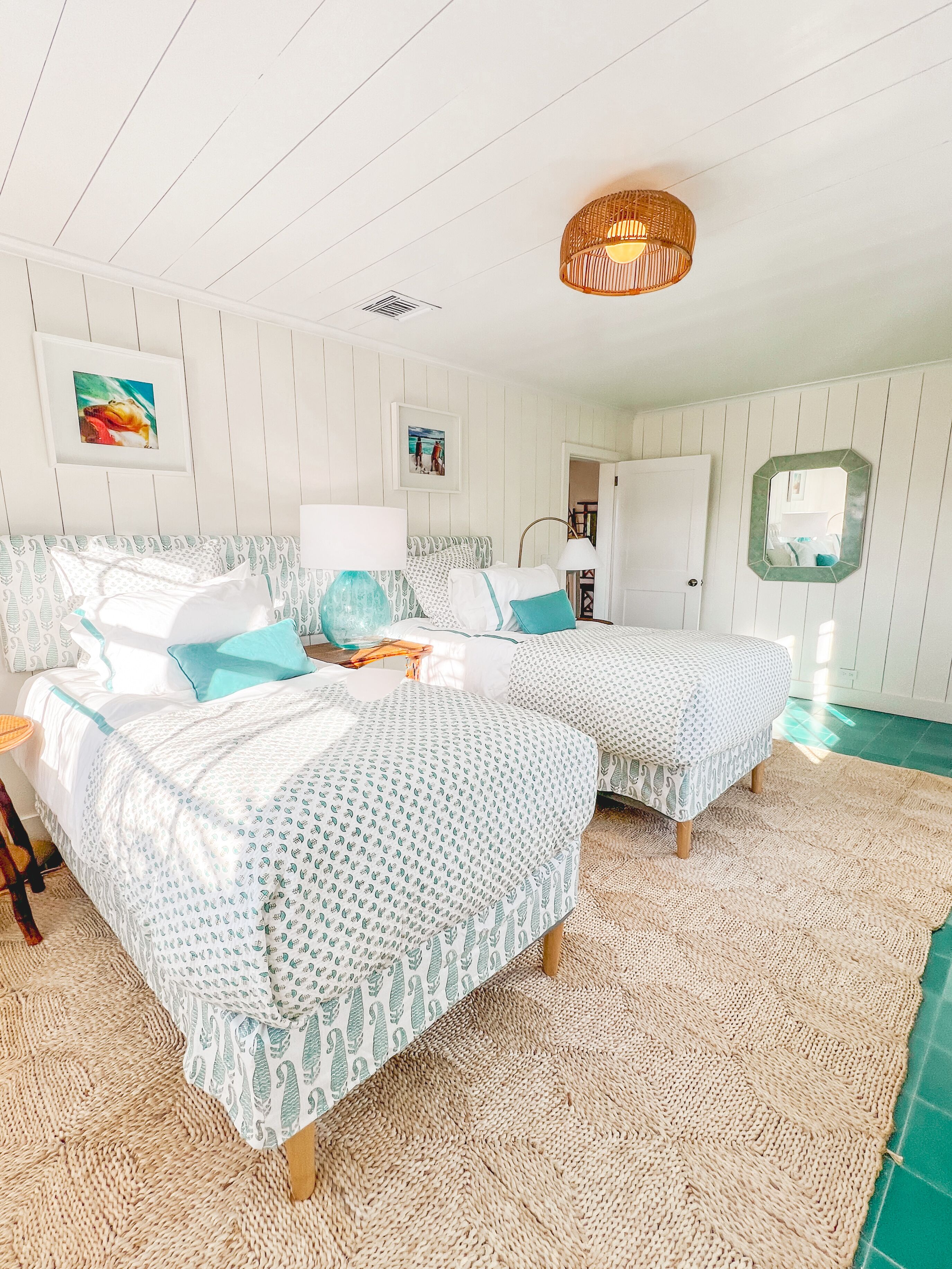 A bedroom with two beds, a woven rug, a hanging light fixture, and a framed picture on the wall.