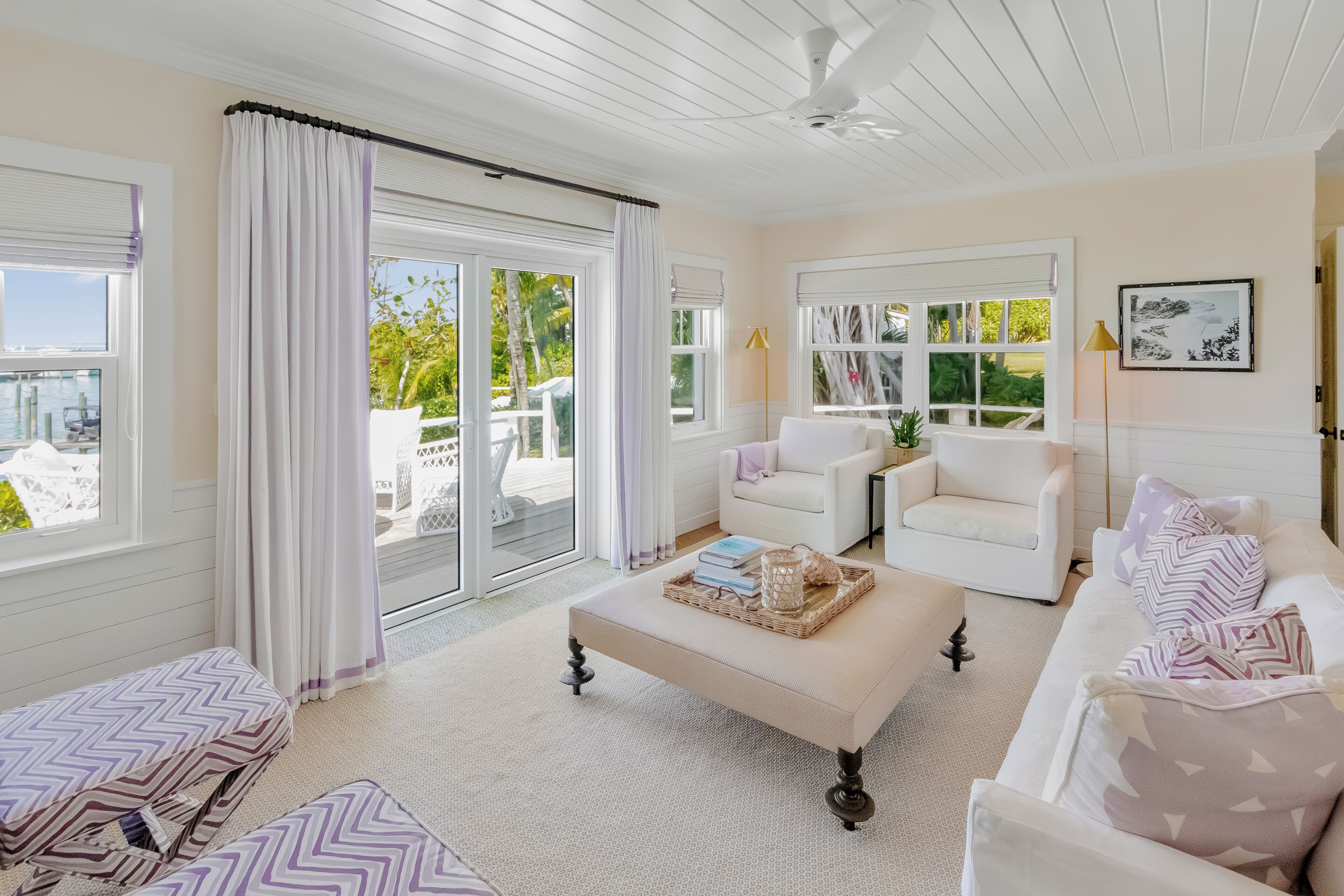 A spacious living room with a large window, a white sofa, a matching armchair, and a coffee table with books and a tray.