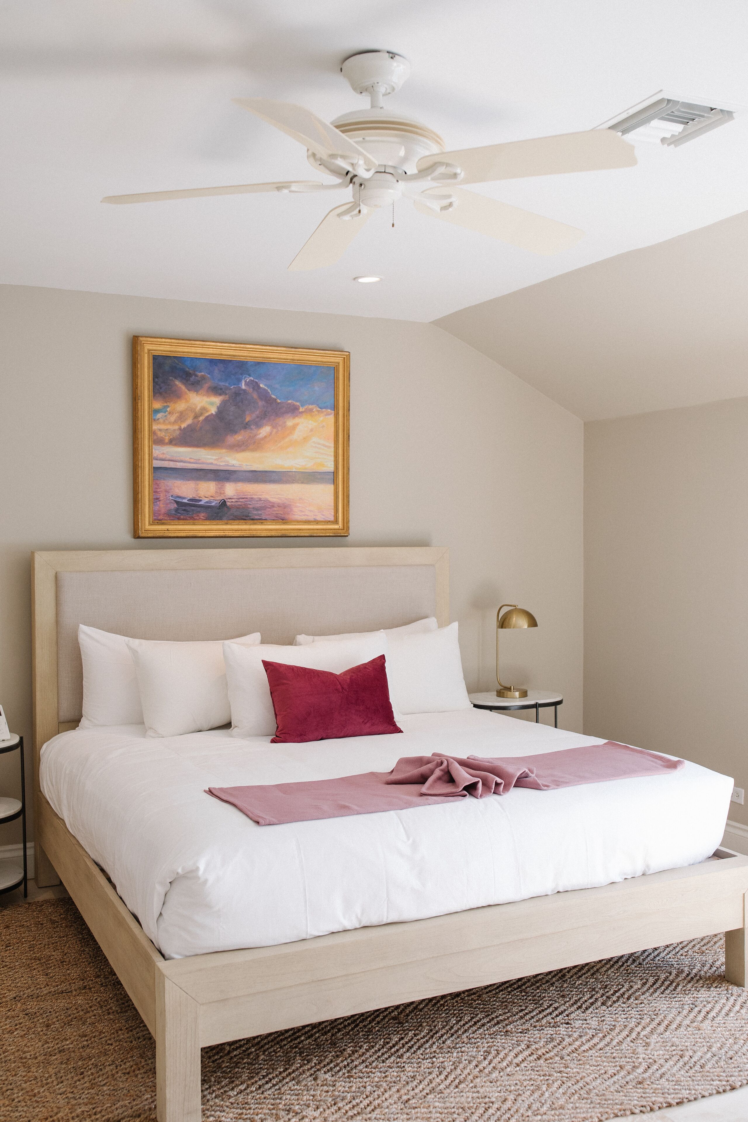 A neatly made bed with white and red bedding, a ceiling fan, a painting of a sunset, and a small bedside table with a lamp.